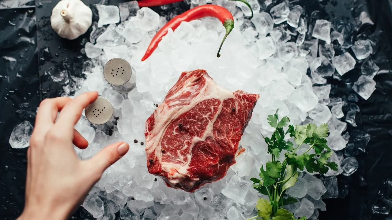 Rohes Rindfleisch-Stück auf zerstoßenem Eis, daneben Knoblauch, Chili und Koriander – Symbolbild zur Kühlung beim Auftauen von Fleisch